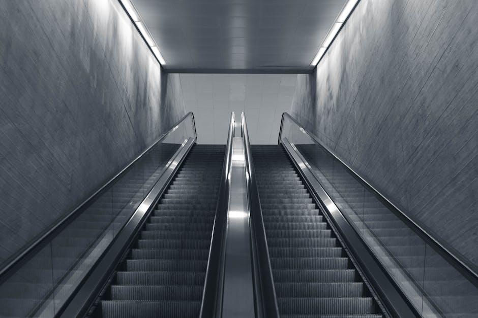 A downward view of a modern escalator with metallic steps and transparent glass side panels, flanked by textured grey walls in a well-lit indoor environment. The escalator is currently stationary, leading to a white ceiling with subtle lighting, highlighting the clean and sleek surface of the moving mechanism. The surrounding surfaces are smooth and free of dust or debris, emphasizing a maintained, hygienic space typical of commercial or residential buildings where surface cleaning and sanitisation are essential. This setting reflects professional cleaning standards suitable for addressing access issues on narrow stairs or escalators, as provided by Maida Vale Carpet Cleaners, specializing in deep cleaning and hygiene maintenance.