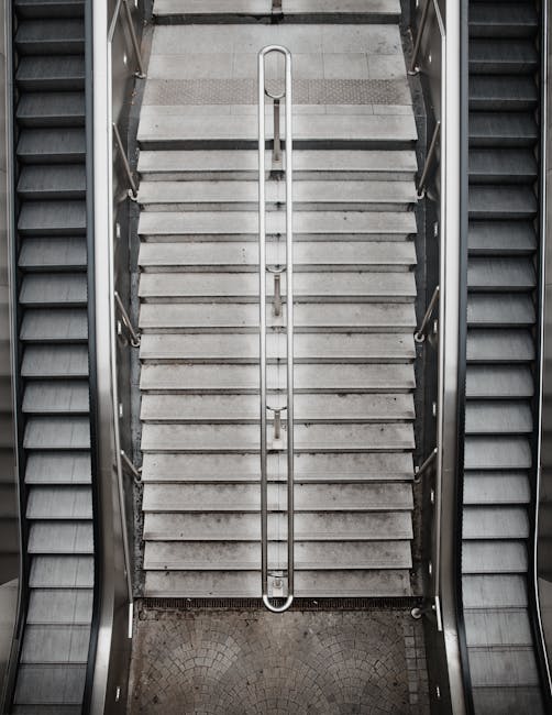 A downward view of a narrow staircase with grey concrete steps featuring textured anti-slip strips. The staircase is flanked by metal railings on both sides, and the lighting is neutral, providing clear visibility of the surfaces. The steps are clean and free of dust or debris, with the surrounding area also in good hygienic condition. The stairs are enclosed by metallic framework, and adjacent to the staircase are other escalators or moving walkways with a similar metal finish. This setting is typical of a public or commercial building interior, such as a shopping centre or office complex, where surface cleaning and maintenance are essential for safety and hygiene. As part of a comprehensive cleaning service, Maida Vale Carpet Cleaners would ensure such areas are thoroughly sanitised to maintain cleanliness and accessibility, especially those with potential access problems as mentioned in the related page. In this image, no cleaning tools or products are visible, emphasizing the importance of regular deep cleaning to preserve surface condition and safety.