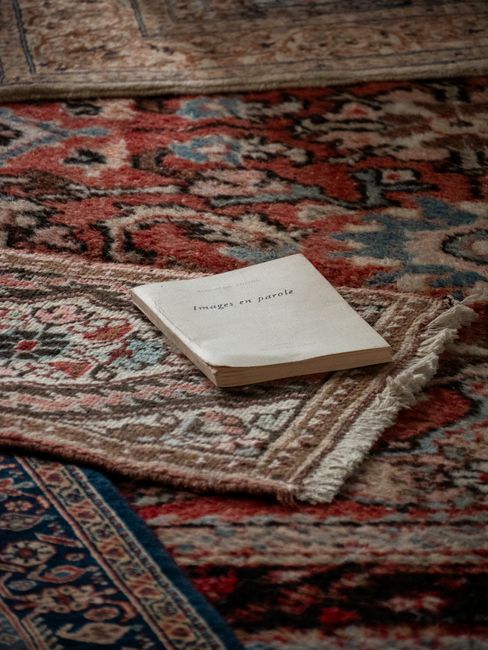 A close-up of a richly patterned, multicolored Oriental rug with intricate floral and geometric designs in shades of red, blue, and beige, laid on a wooden floor. A slightly folded, worn-out hardcover