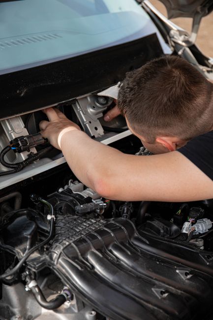 A mechanic inspecting the engine compartment of a car with the hood open, showing various engine components, hoses, and wiring. The mechanic, wearing a black top, is working closely with one hand on a part of the engine, using a tool in the other hand. The engine bay is clean and well-maintained, with visible metal surfaces, plastic covers, and electrical connections. Bright lighting illuminates the area, emphasizing the cleanliness and organization of the engine components. This detailed view highlights surface cleaning and maintenance procedures typical in automotive service, aligned with the importance of surface integrity and systematic deep cleaning in vehicle toolkit and engine sanitation, as part of professional service by Maida Vale Carpet Cleaners.