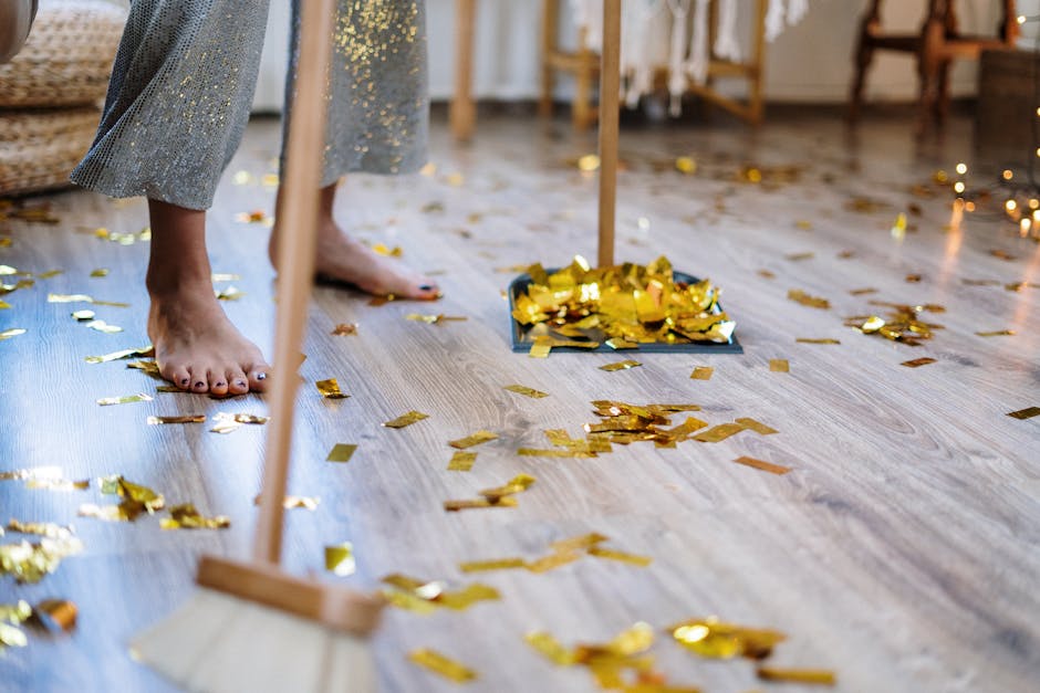 Close-up view of a light wooden floor covered with gold and silver confetti from a celebration, with a person barefoot in grey pants standing near a dustpan filled with confetti, indicating post-party cleaning. The room features warm lighting and background furniture that suggests a living space or dining area, with visible chairs and a table. The scene emphasizes surface cleaning and hygiene, aligned with deep cleaning and sanitisation services provided by Maida Vale Carpet Cleaners, as they restore the cleanliness of residential spaces after events.