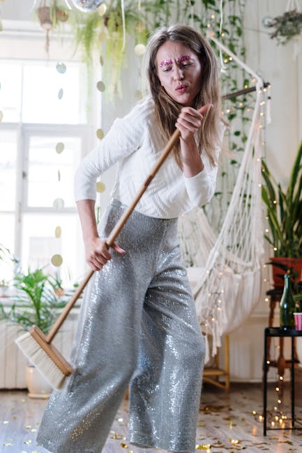 Close-up view of a light wooden floor covered with gold and silver confetti from a celebration, with a person barefoot in grey pants standing near a dustpan filled with confetti, indicating post-party cleaning. The room features warm lighting and background furniture that suggests a living space or dining area, with visible chairs and a table. The scene emphasizes surface cleaning and hygiene, aligned with deep cleaning and sanitisation services provided by Maida Vale Carpet Cleaners, as they restore the cleanliness of residential spaces after events.