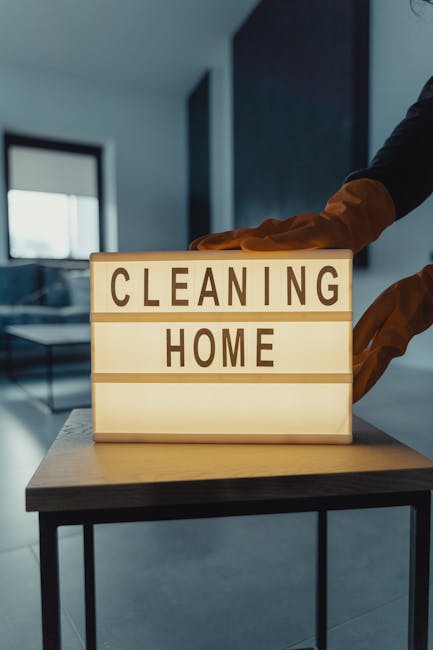 An illuminated lightbox with the words 'CLEANING HOME' prominently displayed in dark letters, placed on a wooden table in a modern, well-lit interior room. The background features a blue wall, a window with natural light streaming in, and a person in a dark jacket with orange gloves reaching towards the lightbox. The scene emphasizes cleanliness and professional domestic cleaning services, consistent with the services offered by Maida Vale Carpet Cleaners, highlighting surface cleaning and hygiene in a contemporary home setting.