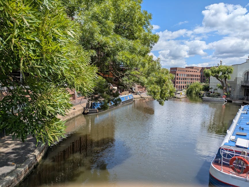 A riverside scene featuring a calm canal with reflective water, lined by lush green trees and shrubs on the left side, and modern residential buildings with large windows and balconies on the right. Several narrowboats are moored along the bank, including a blue boat with a white roof and red lifebuoy. The sky is partly cloudy with bright sunlight illuminating the scene, highlighting the greenery and the clean, well-maintained appearance of the surroundings. This setting showcases a tranquil urban waterway, ideal for surface cleaning and maintenance in residential areas, which Maida Vale Carpet Cleaners specializes in for home and commercial properties.