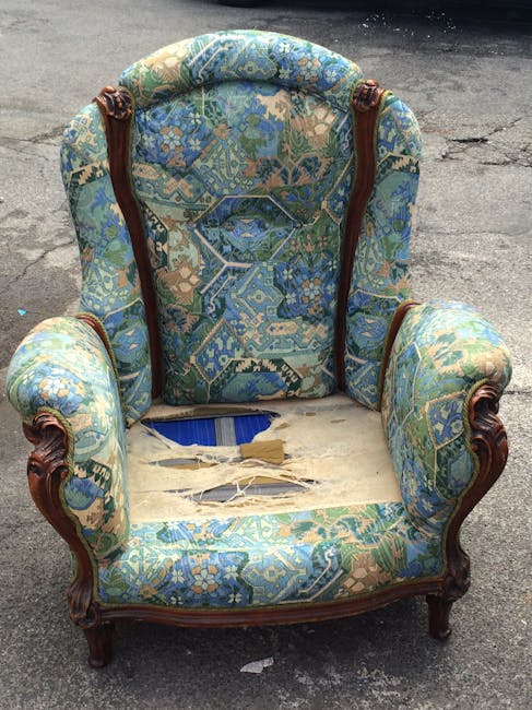 A person wearing a dark long-sleeve shirt is using a handheld vacuum cleaner to deep clean a decorative yellow and white patterned cushion on a dark fabric sofa in a living room. The sofa and cushion are clean and well-maintained, and the room is illuminated with soft lighting. Visible surfaces include the textured fabric of the sofa, the patterned textile of the cushion, and the person's hand gripping the vacuum cleaner. The scene exemplifies professional upholstery surface cleaning and maintenance, consistent with services offered by Maida Vale Carpet Cleaners under their Little Venice upholstery cleaning and stain rescue offerings.