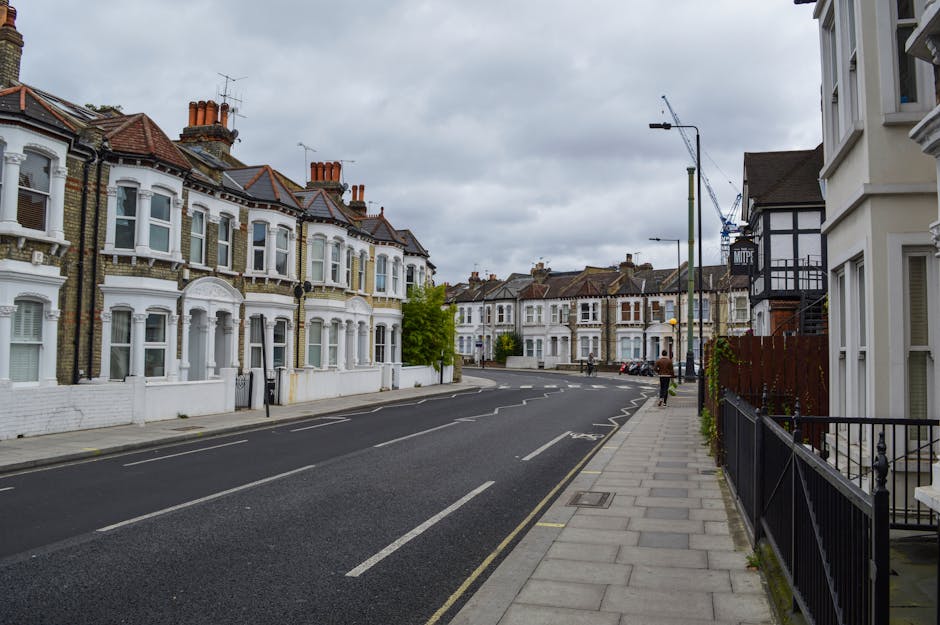 A quiet residential street on Sutherland Avenue showing white Victorian terraced houses with decorative bay windows and red tile roofs. The pavement on the right is clean and lined with a black metal railing, while the asphalt road is marked with white lines and appears freshly maintained. Overhead, the sky is overcast with grey clouds, and there are streetlights and a crane visible in the distance. The scene reflects a tidy, well-kept urban environment consistent with the area served by Maida Vale Carpet Cleaners' domestic and commercial cleaning services, including end of tenancy cleaning.