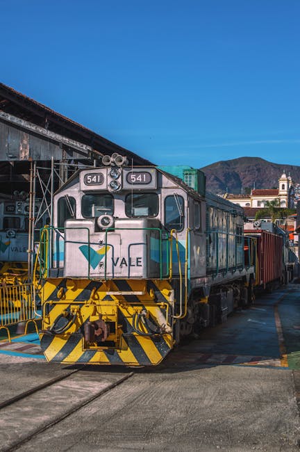 A close-up view of a vintage diesel locomotive situated outdoors on railway tracks, with the front facing directly towards the camera. The train's body is primarily metallic silver with blue and green accents, displaying the number 641 on the front. The foreground shows the yellow and black safety markings on the buffer beam. The surrounding area includes some metal railings and equipment, indicating a train yard or station setting. The background features a clear blue sky, distant hills, and a small building with a clock tower, suggesting a rural or semi-urban environment. Natural daylight illuminates the scene, emphasizing the clean and well-maintained appearance of the locomotive, which is part of a modern fleet operated by Maida Vale Carpet Cleaners for contextually matching surface cleaning themes.