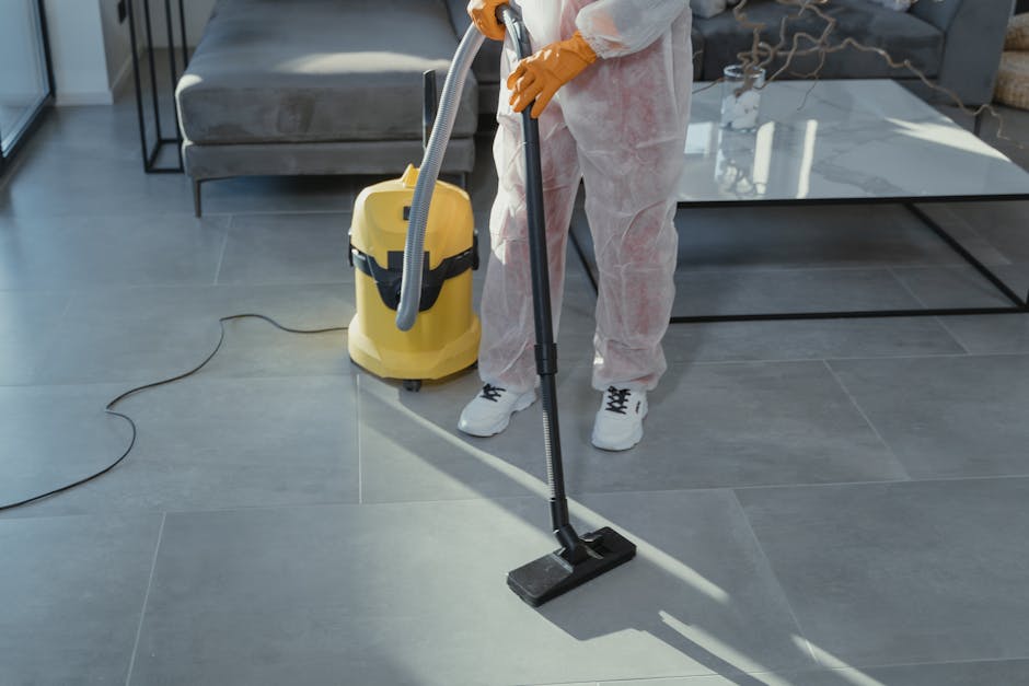 A person dressed in white protective coveralls and orange gloves is using a vacuum cleaner to deep clean a large, grey tiled floor in a modern living room. The vacuum cleaner, which is yellow with a flexible hose, is positioned on the floor and connected to a power outlet via a cord. The individual is holding a black cleaning tool with a long handle, directing it across the tiled surface, which appears clean and free of dust or debris. In the background, there is a grey upholstered sofa, a glass-top coffee table, and decorative items such as a bowl and a plant, illuminated by natural light coming through nearby windows. Maida Vale Carpet Cleaners' service involves surface cleaning and sanitisation to maintain a hygienic living space.