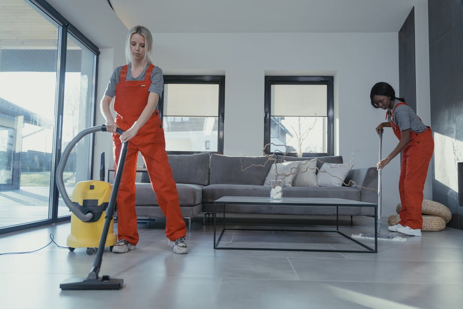 Two professional cleaners from Maida Vale Carpet Cleaners are performing surface cleaning and deep cleaning of a modern living room. The cleaner on the left is vacuuming the grey tiled floor using a yellow canister vacuum cleaner, while the cleaner on the right is using a mop or steam cleaner on the same floor. The living room features a contemporary design with a grey sofa adorned with beige and cream cushions placed against a white wall with large windows allowing natural light to illuminate the space. The room’s surfaces, including a black metal-framed glass coffee table, are visibly clean and tidy, with minimal clutter. Nearby, decorative woven baskets are positioned in the corner, and the natural lighting highlights the neat and hygienic condition of the furniture and flooring. The overall scene depicts professional domestic cleaning focused on sanitisation and maintaining the cleanliness of the living area, aligning with the services offered by Maida Vale Carpet Cleaners for same-day sofa and surface cleaning services on Warwick Avenue.