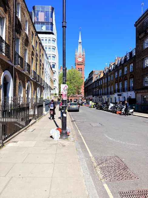 A clear city street in Westminster with historic brick buildings on either side, featuring residential flats with white window frames and small balconies. The sidewalk is made of concrete slabs, with a black metal fence bordering the front yards. A black pole with a street sign and a pedestrian crossing sign stands in the foreground, while a person is walking along the sidewalk, carrying shopping bags. Parked cars, scooters, and motorcycles line the curbside, and in the background, the iconic red brick clock tower of Westminster Council building rises against a bright blue sky. The area appears clean and well-maintained, reflecting regular surface cleaning and city upkeep, linking to hygiene and maintenance principles discussed in Westminster Council waste rules for Maida Vale cleaning firms. Maida Vale Carpet Cleaners emphasizes the importance of professional deep cleaning and sanitisation for maintaining the cleanliness and hygiene of residential and commercial premises in this area.