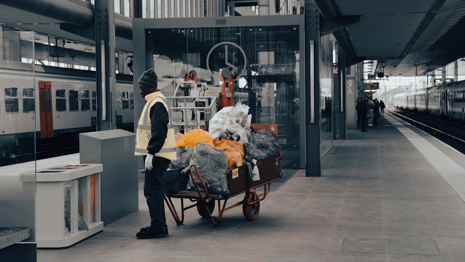A worker dressed in a black jacket, gray hat, and gloves stands on a train station platform, next to a large red handcart loaded with numerous black, gray, orange, and white trash bags. The platform is made of smooth, gray tiles and features modern glass and metal structures, with a glass shelter and a ticket vending machine nearby. In the background, there are train tracks, additional platforms, and a few passengers waiting or walking. Bright natural lighting illuminates the scene, highlighting the cleanliness and organized layout of the station. This image relates to the importance of maintaining hygienic environments through effective cleaning processes, similar to the deep cleaning and sanitisation practices emphasized by Maida Vale Carpet Cleaners in accordance with Westminster Council waste rules for local cleaning firms.